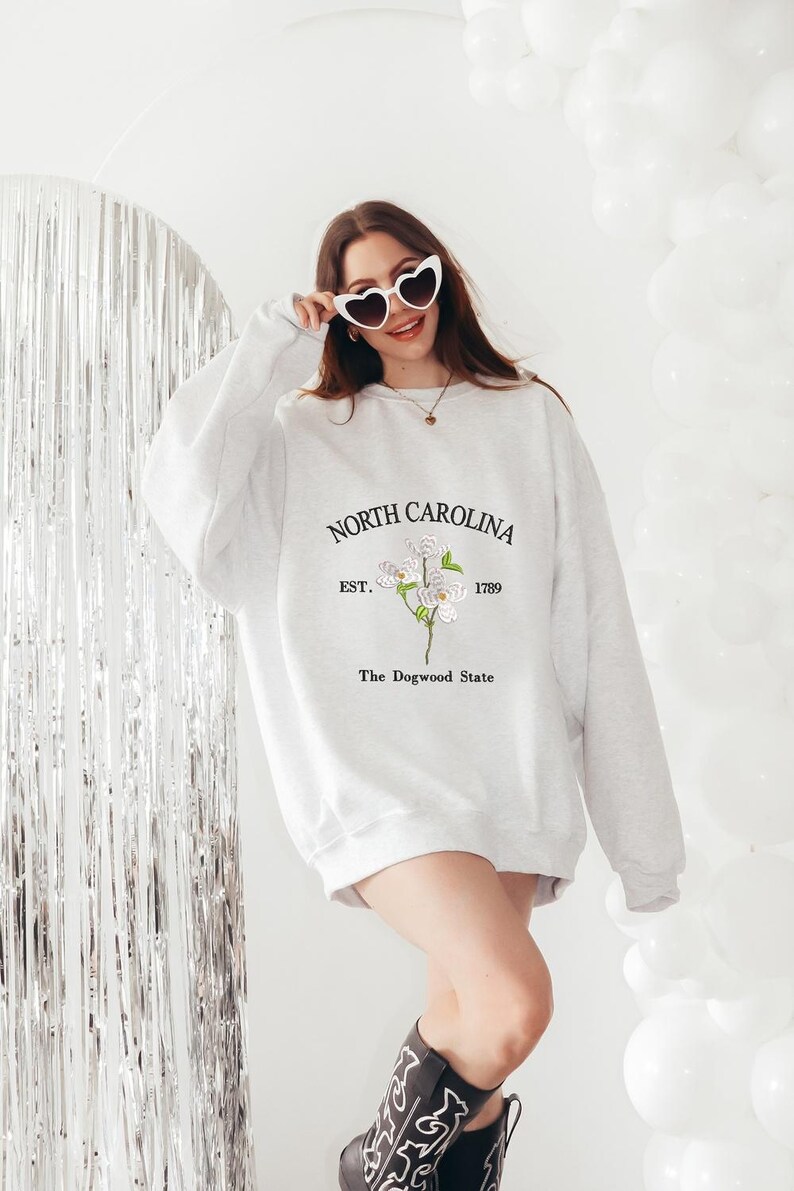 Pu&ograve; includere: Una felpa grigio chiaro con la scritta "NORTH CAROLINA EST. 1789" e una grafica floreale. La felpa ha anche il testo "The Dogwood State". La persona indossa occhiali da sole a forma di cuore e stivali da cowboy.