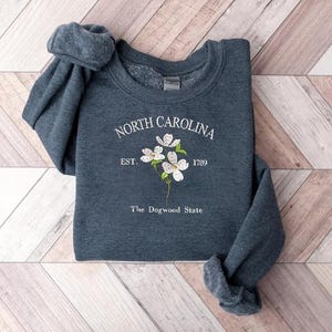 Pu&ograve; includere: Una felpa blu scuro con la scritta "NORTH CAROLINA" in bianco, in un font circolare. Sotto il testo c'&egrave; "EST. 1789" e un disegno floreale. Le parole "The Dogwood State" sono in basso.