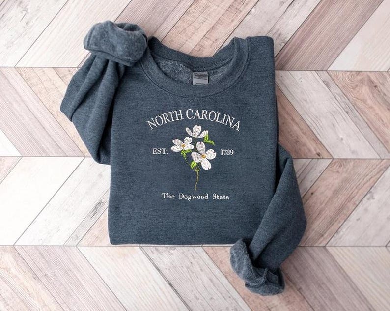 Pu&ograve; includere: Una felpa blu scuro con la scritta "NORTH CAROLINA" in bianco, in un font circolare. Sotto il testo c'&egrave; "EST. 1789" e un disegno floreale. Le parole "The Dogwood State" sono in basso.