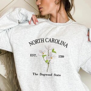 Pu&ograve; includere: Felpa grigio chiaro con la scritta "NORTH CAROLINA" sopra un ricamo di fiore di corniolo e la scritta "EST. 1789" e "The Dogwood State". La felpa ha le maniche lunghe.