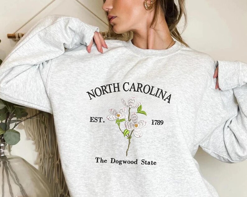 Pu&ograve; includere: Felpa grigio chiaro con la scritta "NORTH CAROLINA" sopra un ricamo di fiore di corniolo e la scritta "EST. 1789" e "The Dogwood State". La felpa ha le maniche lunghe.