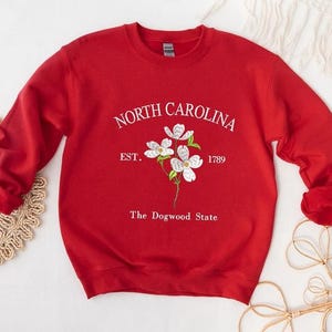 Pu&ograve; includere: Una felpa rossa con la scritta bianca "NORTH CAROLINA" sopra un disegno floreale. Sotto i fiori ci sono le parole "EST. 1789" e "The Dogwood State" in bianco. La felpa ha le maniche lunghe ed &egrave; realizzata in materiale morbido.
