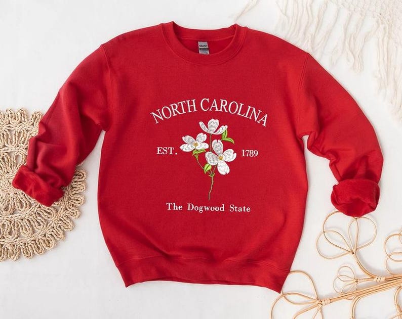 Pu&ograve; includere: Una felpa rossa con la scritta bianca "NORTH CAROLINA" sopra un disegno floreale. Sotto i fiori ci sono le parole "EST. 1789" e "The Dogwood State" in bianco. La felpa ha le maniche lunghe ed &egrave; realizzata in materiale morbido.