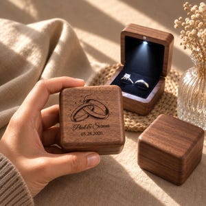 May include: A square dark wood ring box with a hinged lid featuring an engraved design of two interlocking rings, the names Paul & Serena, and the date 05.28.2026. An open matching box displays two silver rings under a small LED light.