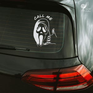 May include: White vinyl decal featuring the Ghostface mask from the Scream movie franchise, with the text "CALL ME" above the mask and a hand giving the shaka sign. The decal is applied to the rear window of a black car.