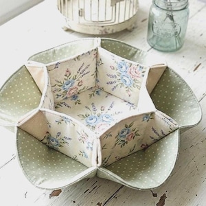 Peut inclure: Un plateau en tissu à six compartiments. L'intérieur présente un tissu crème avec des roses bleues et roses. L'extérieur est vert clair avec des pois blancs. Une cage à oiseaux décorative et un bocal en verre avec de la lavande sont en arrière-plan.