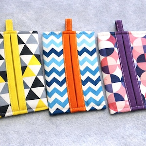 May include: Three rectangular fabric tissue holders. The first has a yellow accent and a geometric pattern of black, white, and yellow triangles. The second has an orange accent and a blue and white zigzag pattern. The third has a purple accent and a pink and purple circle pattern.