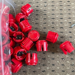 Könnte beinhalten: Nahaufnahme eines Stapels roter Arcade-Knöpfe in einer durchsichtigen Plastiktüte. Die Knöpfe sind in zufälliger Reihenfolge angeordnet und einige sind teilweise sichtbar. Die Knöpfe sind aus Kunststoff und haben eine runde Form mit einer flachen Oberseite.