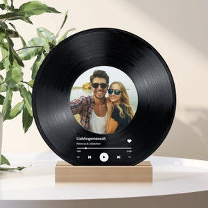 Può includere: Un disco in vinile nero con una foto di una coppia con occhiali da sole. Il disco è su un supporto in legno con un'interfaccia di lettore musicale che dice "Lieblingsmensch" e "Rebecca & Sebastian".