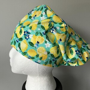 May include: A scrub cap with a bright lemon print on a teal backdrop. The design features yellow lemons, white flowers, and blue details. The cap is displayed on a white mannequin head.