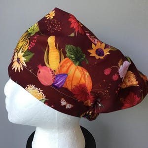 May include: A burgundy scrub hat with a fall-themed print featuring pumpkins, corn, pomegranates, and sunflowers. The hat has a tie at the back.