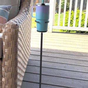 May include: A tall, thin black metal stand with an X-shaped base holds a teal and purple ombre tumbler with a handle and a matching teal straw. The cup is secured in a circular holder at the top of the vertical pole, providing a stable surface.