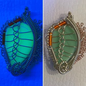 Könnte beinhalten: Ein grüner Glasanhänger mit einem silbernen Drahtkäfig-Design. Der Anhänger leuchtet unter Schwarzlicht in einem leuchtenden Grün.