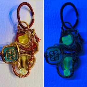 Könnte beinhalten: Ein Anhänger aus Draht und Glas. Der Anhänger besteht aus Kupferdraht und hat ein grünes, gelbes und klares Glasdesign. Der Anhänger wird sowohl in normalem als auch in UV-Licht gezeigt.