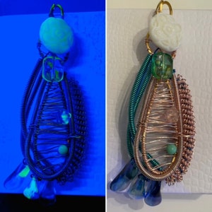 Könnte beinhalten: Ein Anhänger aus Kupferdraht, Perlen und Glas. Der Anhänger hat die Form einer Träne und ist mit einer weißen muschelförmigen Perle oben versehen. Der Anhänger leuchtet unter Schwarzlicht grün.