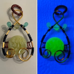 Könnte beinhalten: Ein Anhänger aus goldfarbenem Draht mit einem grünen Glasknopf in der Mitte. Der Anhänger ist mit kleinen blauen Glasperlen und einer Schlaufe aus Kupferdraht oben verziert. Der Anhänger leuchtet unter Schwarzlicht grün.