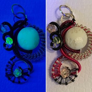 May include: A black and red wire wrapped keychain with a white button and a green and white bead. The keychain is photographed under a blue light.