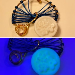 Könnte beinhalten: Ein handgefertigtes Drahtkunstwerk in Blau und Gold. Das Design umfasst einen kreisförmigen Rahmen mit blauem Draht, eine goldene Spirale und eine weiße, kreisförmige Seife mit floralem Muster. Das untere Bild zeigt die Seife leuchtend.