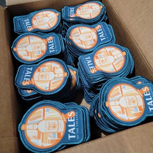 May include: A box filled with blue and orange fabric patches. Each patch features a bottle of Tito's Vodka and the text "Tales of the Cocktail 2024".