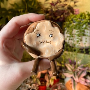 May include: A hand-painted ceramic figurine of a brown and white donut with a smiling face and brown legs. The donut is sitting on a hand.