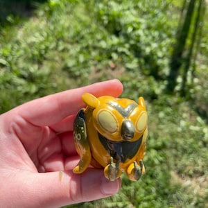 May include: A small, hand-painted ceramic owl figurine with yellow and brown paint. The owl has large, round eyes and a wide beak.