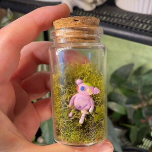 May include: A small, pink, hand-painted clay figure with colorful details sits in a glass jar filled with green moss. The jar has a cork stopper.