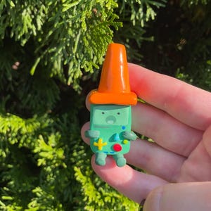 May include: A green figurine of a character with an orange hat. The figurine has a rectangular body with a face and arms. It has a yellow cross, a red circle, and blue dots on its body. The figurine is held in front of a green leafy background.