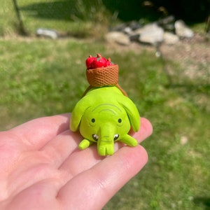 May include: A green elephant figurine with a basket of red apples on its head. The elephant has a small trunk and big eyes. The figurine is made of clay and is about 4 inches tall.