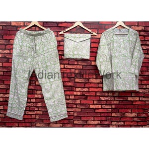 May include: A three-piece pajama set in a light green floral pattern. The set includes pants, a long-sleeved top, and a matching pouch. The garments are displayed on wooden hangers against a red brick wall. The text "indian textile work" is visible.