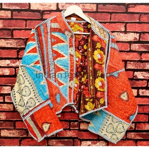 May include: A quilted jacket featuring a patchwork design with various patterns including floral motifs, geometric shapes, and paisley prints in vibrant shades of orange, blue, yellow, and cream, finished with black and white striped trim edges.