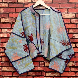 May include: A light blue quilted jacket featuring a vibrant floral pattern in shades of red, orange, and green. The garment has a mandarin collar, wide sleeves, and is finished with dark purple piping along the edges and seams throughout the piece.