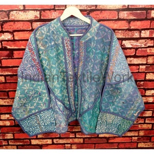 Könnte beinhalten: Blau-türkisfarbene Steppjacke mit floralem Muster. Die Jacke hat ein kragenloses Design und lange Ärmel. Die Jacke hängt an einem weißen Kleiderbügel vor einer Backsteinmauer.
