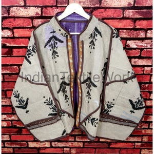 Op de afbeelding: Beige jas met zwarte bladeren en bruine randen. De jas heeft een paarse voering en de tekst "Indian Textile Work". De jas hangt aan een witte hanger tegen een rode bakstenen muur.