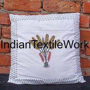 Puede incluir: Funda de cojín de algodón blanco con un borde cosido negro. La funda de cojín presenta un diseño estampado a mano de una planta verde estilizada con hojas amarillas y flores rojas. El texto "Indian TextileWork" está impreso en negro en la funda de cojín.