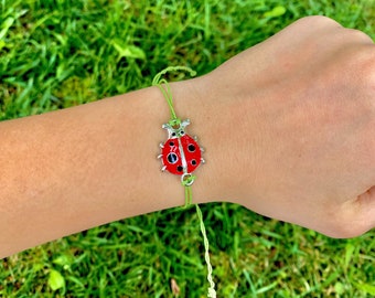 Puede incluir: Un brazalete de cuerda verde con un dije de mariquita plateado. La mariquita es roja con manchas negras y tiene un contorno plateado.