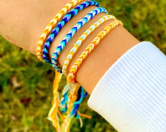 Puede incluir: Un conjunto de cinco pulseras de la amistad en amarillo, azul y blanco. Las pulseras están tejidas en un patrón de chevron y tienen borlas largas.