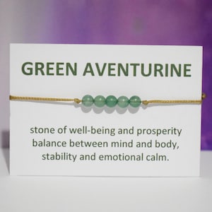 Puede incluir: Un brazalete de aventurina verde con un cordón dorado. La tarjeta dice "GREEN AVENTURINE piedra de bienestar y prosperidad equilibrio entre la mente y el cuerpo, estabilidad y calma emocional."
