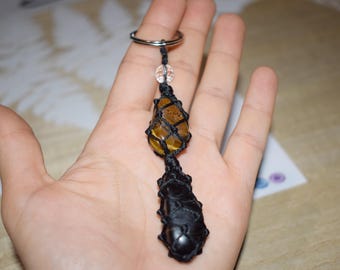 May include: A black cord knotted around a tiger's eye stone and a black stone, with a silver key ring attached.