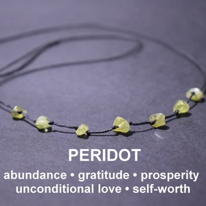 May include: A necklace made with a black cord and small, light green peridot stones. The necklace is on a grey surface. The text "PERIDOT" is below the necklace, and the text "abundance · gratitude · prosperity unconditional love · self-worth" is below that.