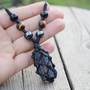 Op de afbeelding: Een zwarte macramé ketting met een donkere, rechthoekige stenen hanger. De ketting bevat zwarte, bruine en zilverkleurige kralen. De hanger is omhuld in een geweven net, gepresenteerd op een houten oppervlak.