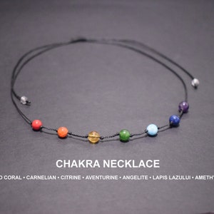 May include: A black cord necklace with seven coloured gemstone beads representing the chakras. The beads are red, orange, yellow, green, light blue, dark blue, and purple. The necklace is labelled "Chakra Necklace" with the names of the stones.