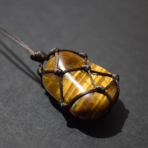 Può includere: Una collana con cordoncino marrone con un ciondolo in occhio di tigre dorato e marrone avvolto in un nodo macramé.