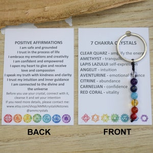 May include: A white card with the text "POSITIVE AFFIRMATIONS" and a list of affirmations.  The card is next to a white card with the text "7 CHAKRA CRYSTALS" and a list of crystals and their meanings.  A silver key chain with seven colored beads is attached to the card.
