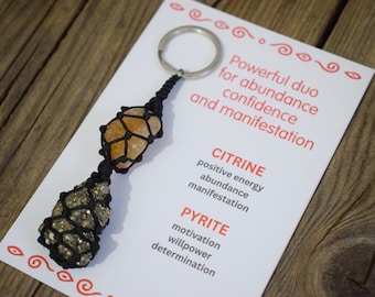 May include: A keychain featuring citrine and pyrite stones, encased in black macrame. The citrine is a warm orange color, while the pyrite has a metallic sheen. A card with red text reads "Powerful duo for abundance, confidence, and manifestation."