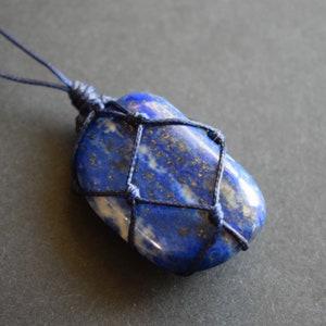 Puede incluir: Un colgante de lapislázuli azul oscuro envuelto en un cordón azul oscuro. La piedra tiene un patrón de mármol con motas blancas y doradas.