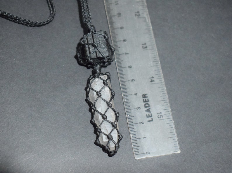 May include: A black cord necklace with a clear crystal pendant wrapped in black macrame. The crystal is about 5 cm long and is next to a ruler for size reference.
