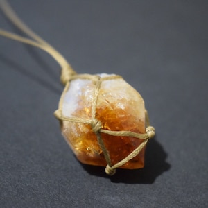 Può includere: Una collana con un ciondolo in cristallo di citrino dorato avvolto in un nodo macramé. Il cristallo è di colore arancio chiaro e ha una texture naturale e ruvida. Il nodo macramé è fatto di cordoncino marrone ed è annodato in un design semplice ed elegante.