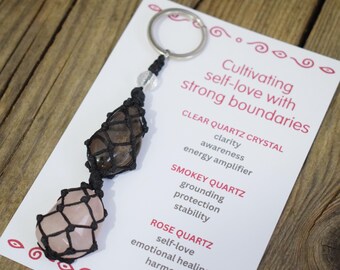 May include: A keychain featuring three crystals: clear quartz, smokey quartz, and rose quartz, each encased in a black macrame net. The keychain includes a silver ring and a clear bead. A card with text about self-love and crystal properties is in the background.