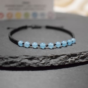 Peut inclure: Un bracelet macramé noir orné de sept perles rondes bleu clair. Les perles sont espacées uniformément le long du cordon du bracelet. Le bracelet est présenté sur une surface gris foncé.