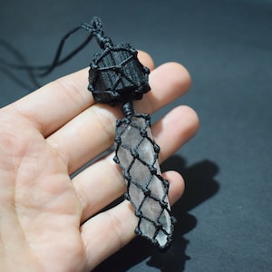 May include: A clear crystal point wrapped in black macrame cord, hanging from a black cord necklace.